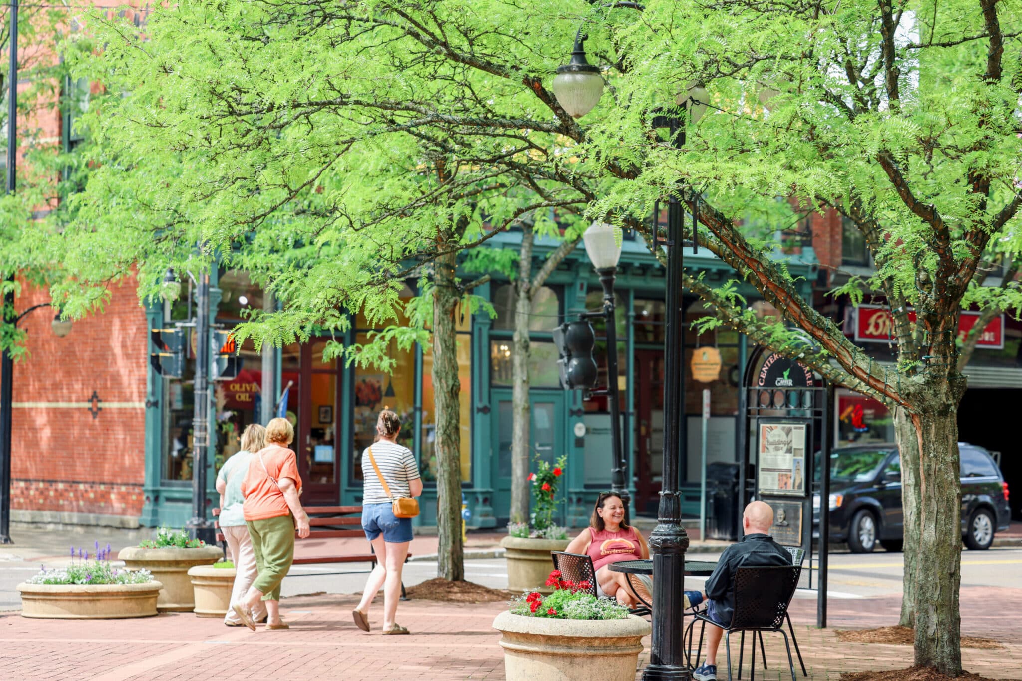 Pedestrians strolling a tree-lined downtown block past shops and cafe seating
